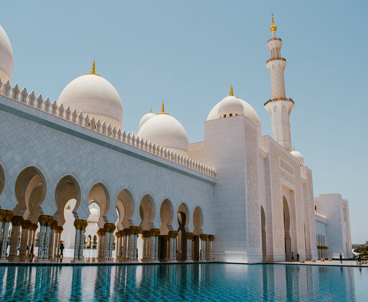The arcade of the Sheikh Zayed Grand Mosque in Abu Dhabi, its white marble domes and minaret above a turquoise reflecting pool