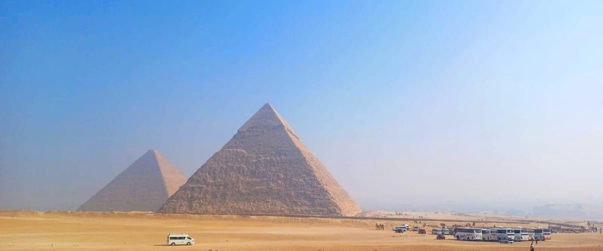 The pyramids of Khafre and Menkaure rising out of the desert at Giza, coaches and camels small along the plateau below them