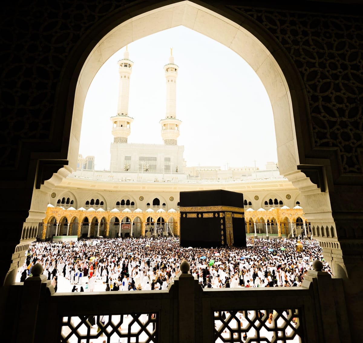 The Kaaba at the centre of Masjid al-Haram seen through a tall arch of the mosque, thousands of pilgrims filling the courtyard around it